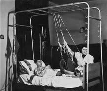A patient being encouraged by a nurse to use the hoists above his bed to pull himself up.