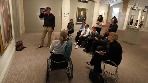 A group of people cluster around a painting hanging on a wall in an art gallery. Most are seated but three are standing. One person is talking, and the others are listening. They are all looking at the painting, which we cannot see.