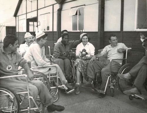 Black and white photo of Christine and athletes outside the huts at Stoke Mandeville.