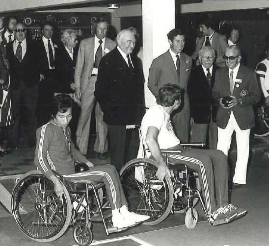 Prince Charles, Sir Ludwig Guttman and Jack Sutherland in the Indoor Bowls Centre at the 1973 International Games with two wheelchair bowlers and other officials behind them.